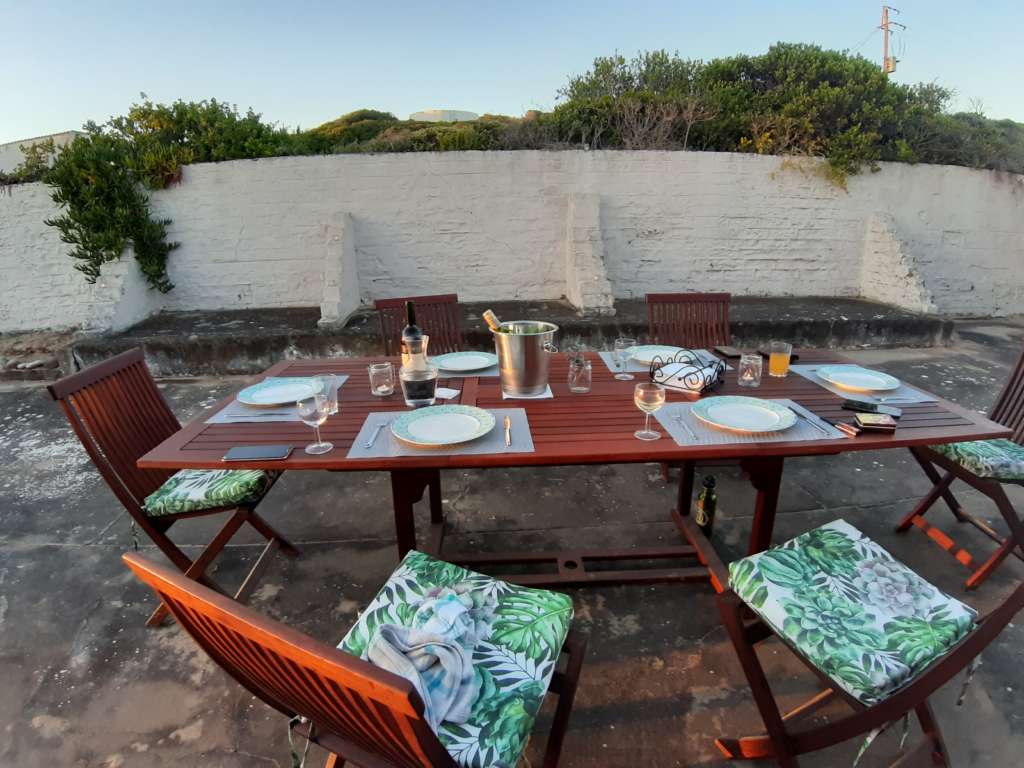 A teak outdoor dining table with place settings, chairs, and cushions, set up near the braai at Fonteintjies.
