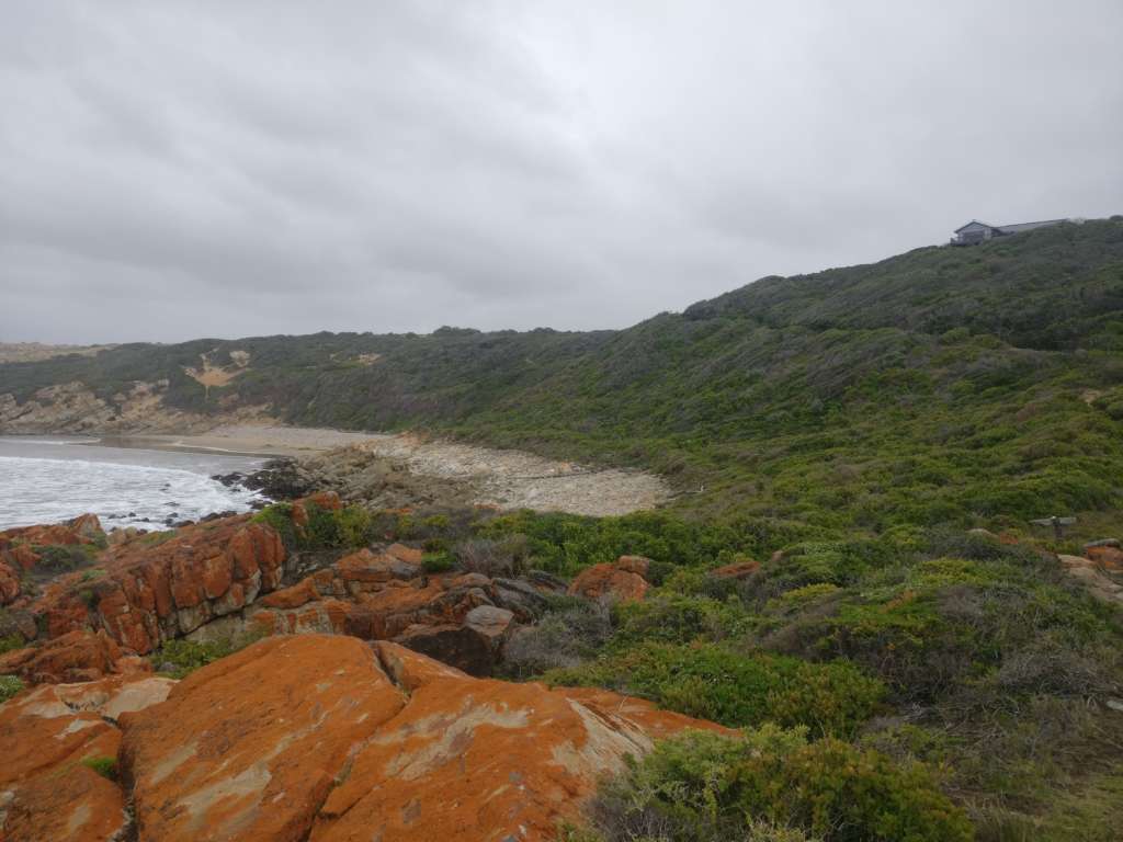 A rocky sea shore. A sandy beack is visible in the distance. The house at Fonteintjies is on the horizon at the right.