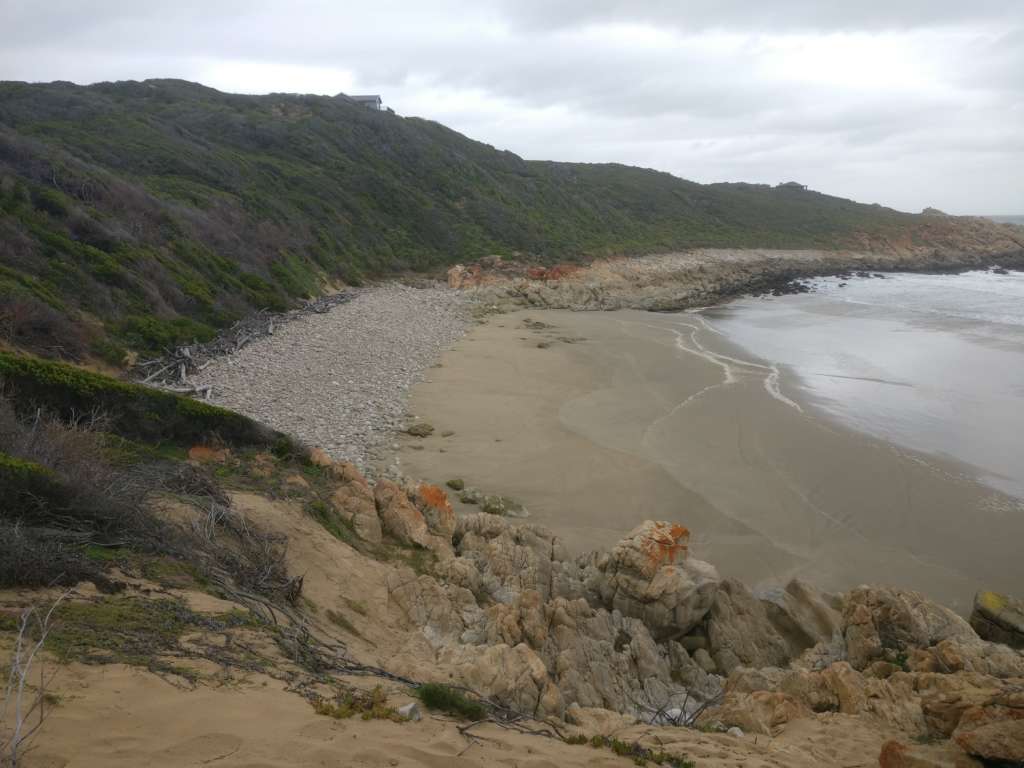 Blick hinunter auf einen kleinen Strand. Das Meer ist rechts. In der Ferne ist ein Hügel mit grüner Vegetation bedeckt. Das Haus und das Deck von Fonteintjies sind am Horizont sichtbar. Ein Nachbarhaus befindet sich in der Ferne rechts.