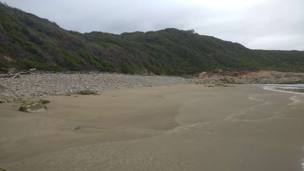 A sandy beach in the foreground, with the ocean behind the photographer. In the middle distance is a rocky part of the beach, with a hill beyond. On the horizon at the top of the hill is the Fonteintjies house.