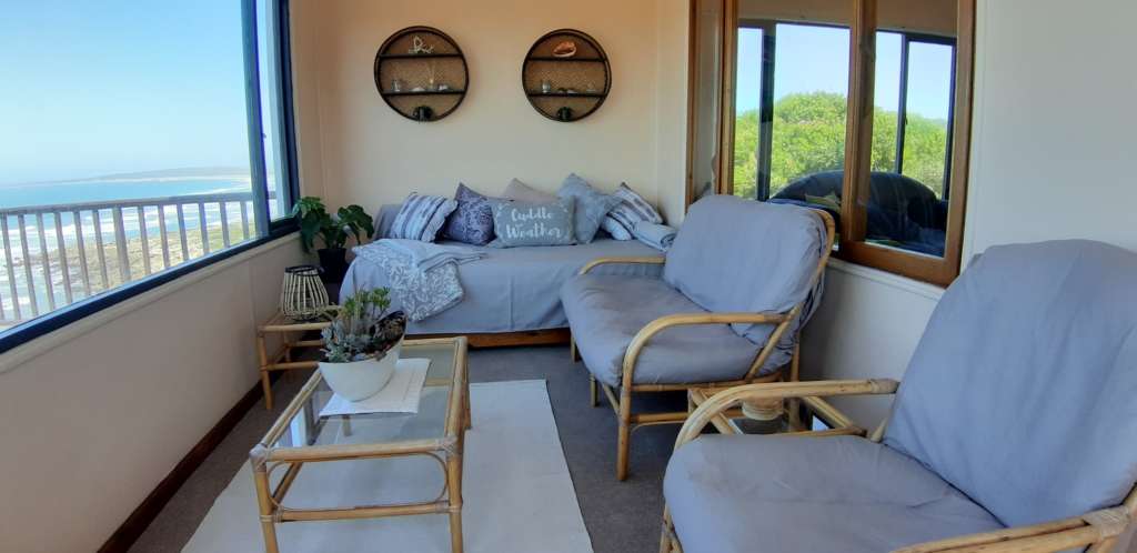 The sun room at Fonteintjies. The foreground shows cane furniture with cushions covered in grey fabric. In the centre of the image are two round decorative shelves with shells, plants and small ornaments. Below the shelves is a day bed with several cushions. "Cuddle Weather" is written on a cushion. The ocean is visible through a window to the left.