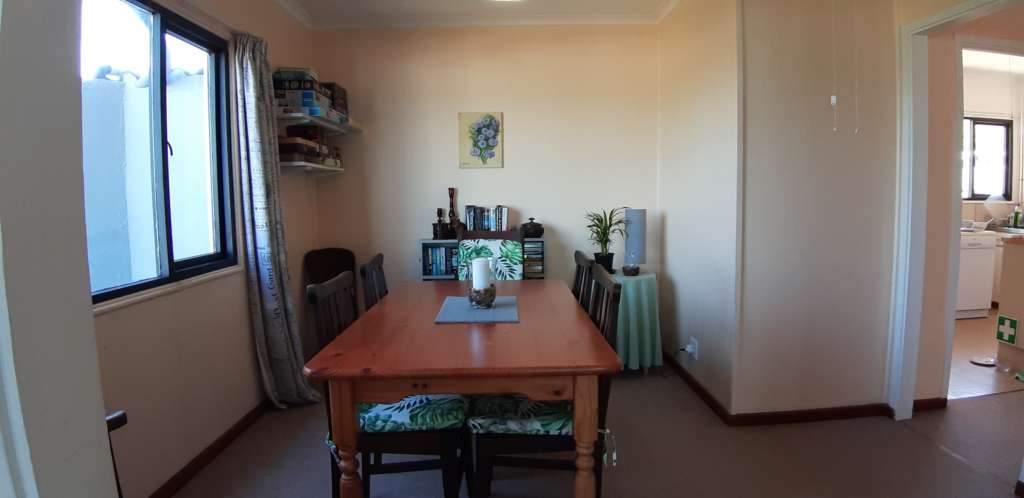 The dining room at Fonteintjies. In the foreground is a wooden dining table with wooden chairs. There is a candle in a glass bowl on the table. Against the wall in the centre is a bookcase, with a painting on the wall above it. In the corner to the left are some shelves with games and puzzles. A round table with a lamp and pot plant are in the corner to the right.