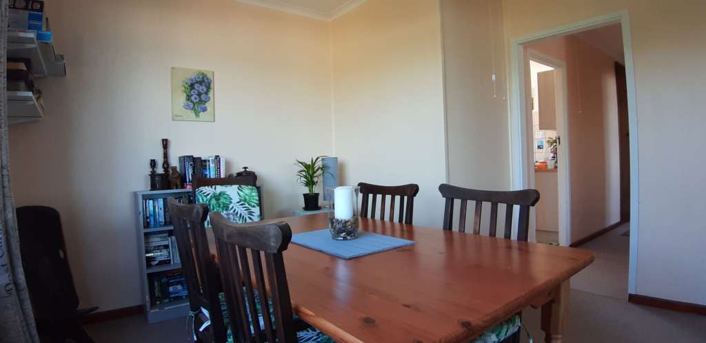 The dining room at Fonteintjies, with the kitchen beyond. In the foreground is a wooden dining table with wooden chairs. There is a candle in a glass bowl on the table. Against the wall in the centre left is a bookcase, with a painting on the wall above it. Just visible to the fat left are some shelves with games and puzzles. Through a door to the right, part of the kitchen is visible.