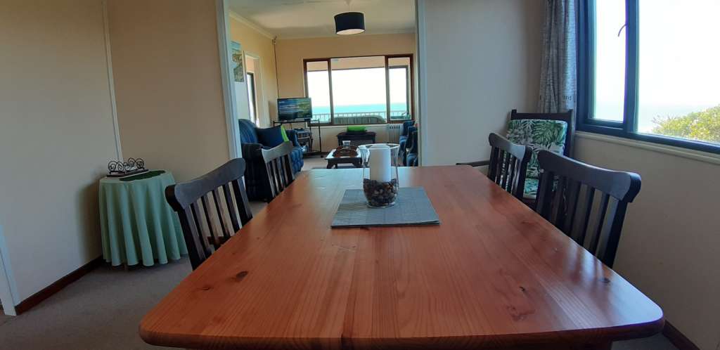 The dining room at Fonteintjies, with the lounge beyond, and the ocean visible in the distance through the sun room. In the foreground is a wooden dining table with wooden chairs. There is a candle in a glass bowl on the table.