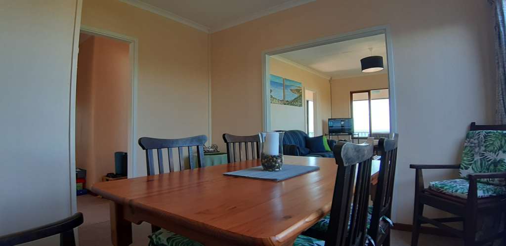 The dining room at Fonteintjies. In the centre is a wooden table with four wooden chairs. A fifth chair is partially visible to the left, and a sixth chair is in a corner to the right. The kitchen is through the door to the left. The lounge is in the background.