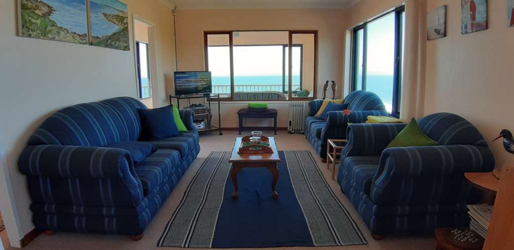 The lounge at Fonteintjies. Large blue striped chairs and couches are against the walls to the left and right. On the left hand wall is a pair of oil paintings depicting the sea as viewed from the deck. A blue and white rug is on the floor. A coffee table is in the centre of the room, with a tray on top of it. A TV is in the far left corner. Also to the far left is the doorway that leads to the deck. In the centre distance, the ocean is visible through a window to the sun room. A sculpture of an oystercatcher bird is partially visible in extreme right of the photo.