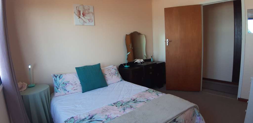 Bedroom 2 at Fonteintjies. In the centre is a double bed. On the wall above the bed is a picture of a flower. In the far right corner is a dressing table with a mirror, a large shell, and a lamp. The entrance door to the right of the picture is open, leading to a passageway.