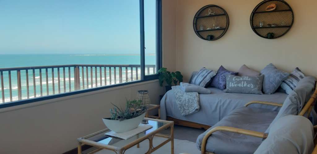 The sun room at Fonteintjies. To the left is a large window, with a view of the ocean. In the centre of the sun room is a table with a display of succulents in a white oval planter. Around the edges of the room are cane chairs and couches with cushions covered in grey, and at the far left is a day bed with blankets and cushions. On the wall above the day bed are two round decorative shelves, with shells, plants and other items.