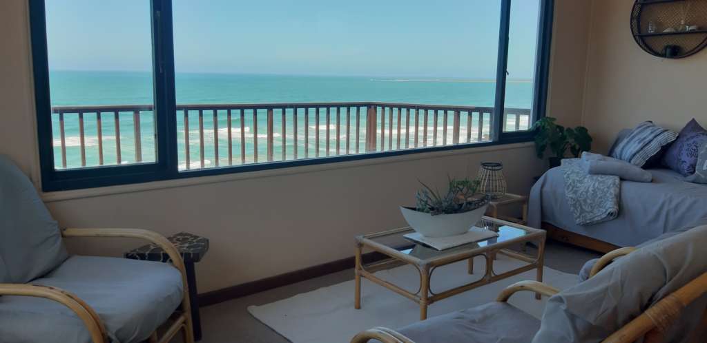 The sun room at Fonteintjies. Dominating the photo is a large window, with a view of the ocean. In the centre of the sun room is a table with a display of succulents in a white oval planter. Around the edges of the room are cane chairs and couches with cushions covered in grey, and at the far left is a day bed with blankets and cushions.