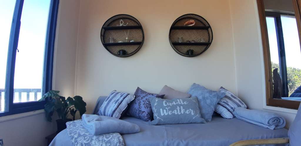 The sun room at Fonteintjies. In the centre of the image are two round decorative shelves with shells, plants and small ornaments. Below the shelves is a day bed with several cushions. "Cuddle Weather" is written on a cushion.