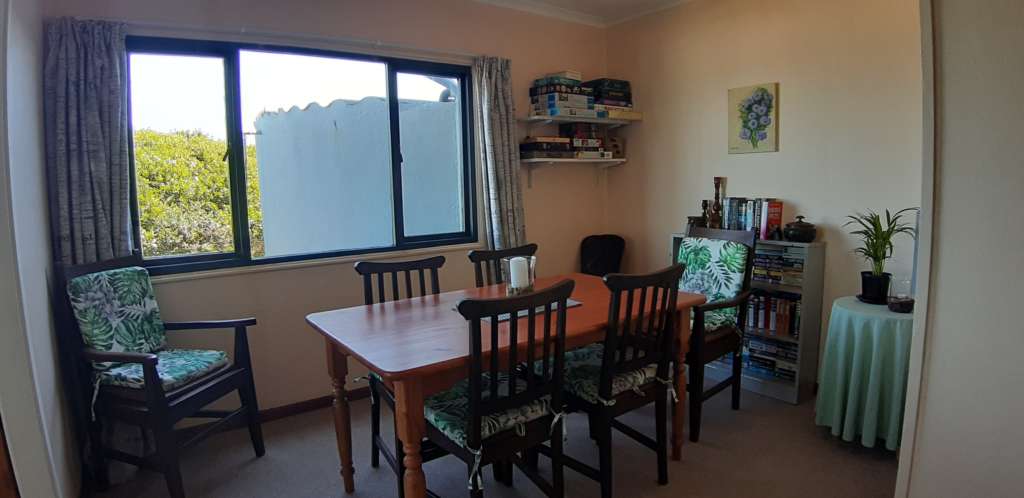 The dining room at Fonteintjies. In the centre is a wooden table with five wooden chairs and cushions. A sixth matching chair is in the left corner. There is a candle in a glass bowl on the table. A large window on the left hand wall shows a view of vegetation and a water tank outside. In the left rear corner are shelves with games and puzzles. On the wall to the right rear is a bookcase full of books. On the wall above the bookcase is a painting of a bunch of flowers. In the far right corner is a table with a lamp and pot plant.