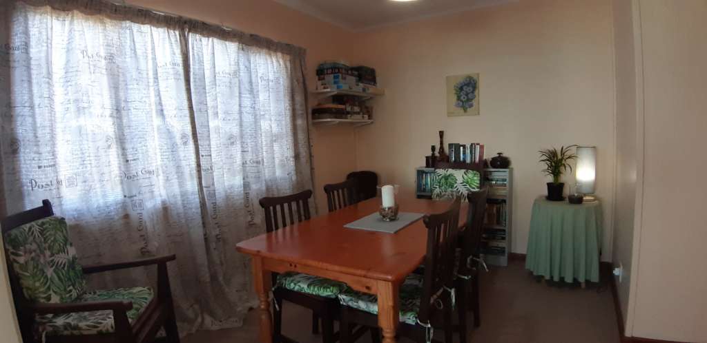 The dining room at Fonteintjies. In the centre is a wooden table with five wooden chairs and cushions. A sixth matching chair is in the near left corner. There is a candle in a glass bowl on the table. A large window on the left hand wall is covered by curtains. In the left rear corner are shelves with games and puzzles. On the wall in the centre rear is a bookcase full of books. On the wall above the bookcase is a painting of a bunch of flowers. In the right rear corner is a table with a lamp and pot plant.