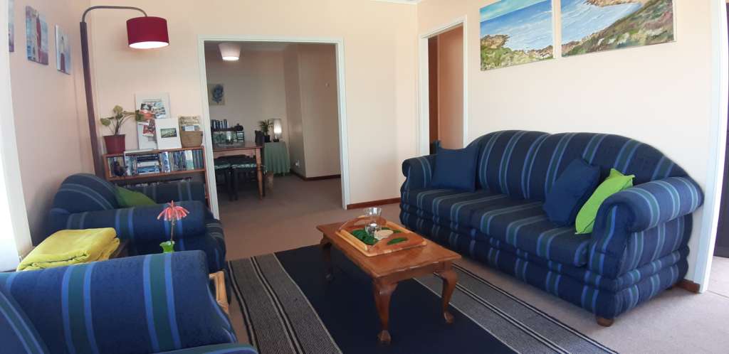The lounge at Fonteintjies. In the centre is a coffee table with a tray on it. To the right is a blue striped three-seater couch. Above the couch are a pair of oil paintings depicting the sea view as seen from the deck. Along the left hand wall are a blue striped two-seater couch, only partially visible, and a matching chair. In the left corner beyond the chair is a standard lamp and a bookcase full of DVDs. On top of the bookcase is a plant and some pamphlets. The dining room is visible through an opening in the wall to the centre rear. Just out of sight to the right is the door to the deck.