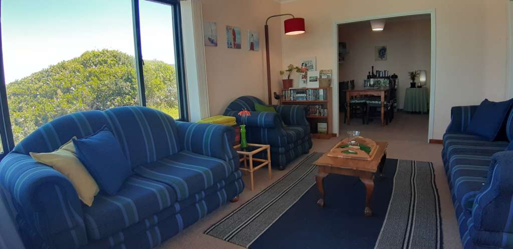 The lounge at Fonteintjies. In the left foreground is a blue striped two-seater couch, with a matching char to its right. A matching three-seater couch is on the right hand wall. In the left corner beyond the chair is a standard lamp and a bookcase full of DVDs. On top of the bookcase is a plant and some pamphlets. The dining room is visible through an opening in the wall to the centre rear.