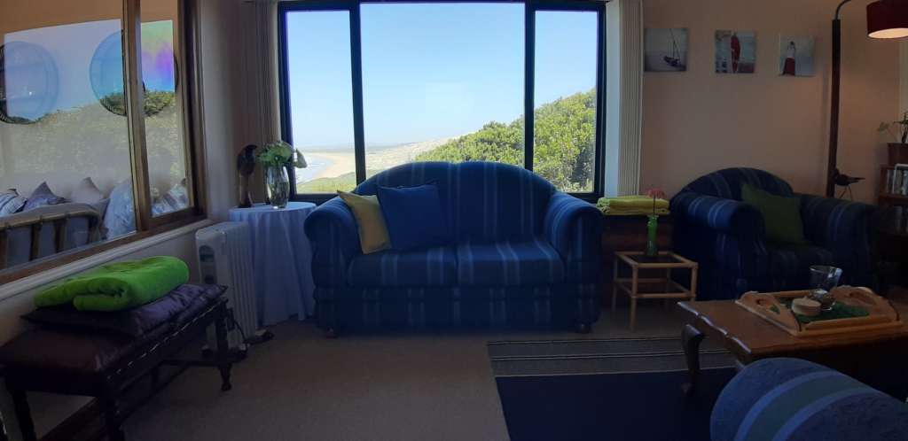 The lounge at Fonteintjies. Behind a large blue striped couch is a window with the ocean visible in the distance. To the left is a window that separates the lounge from the sun room.