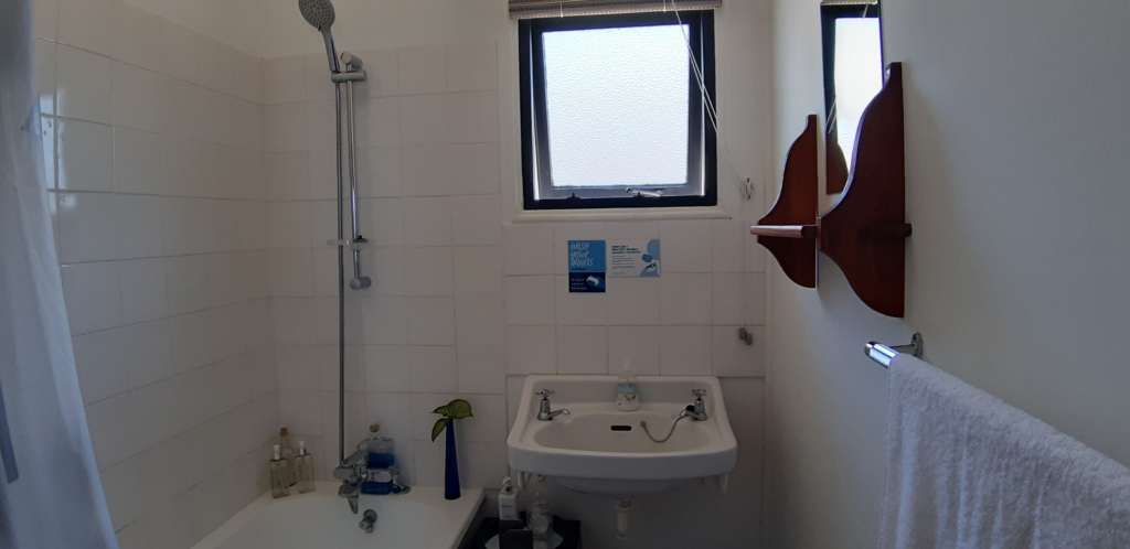 The bathroom at Fonteintjies. In the centre is a hand basin with a window above it. To the left is a bathtub. A shower head is mounted on a rail above the bathtub. On the right hand wall is a mirror with a shelf below it.