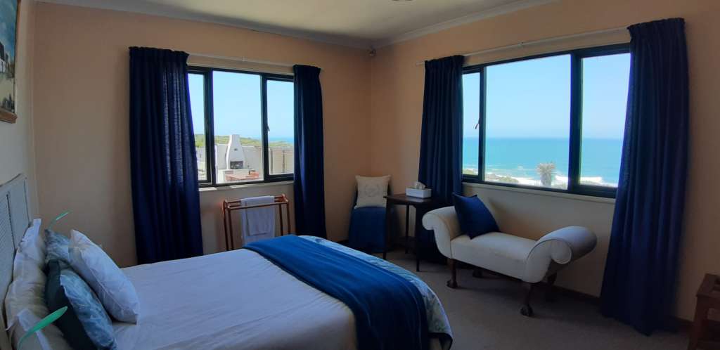 Bedroom 1 at Fonteintjies, seen from near the door. There is a double bed in the foreground. The walls to the far left and centre both have large windows. Below the centre window is a large bedroom seat. In the far corner of the room near the centre of the picture, is a small table. The ocean is visible through the window to the centre right, and the braai and patio are visible through the other window to the left.