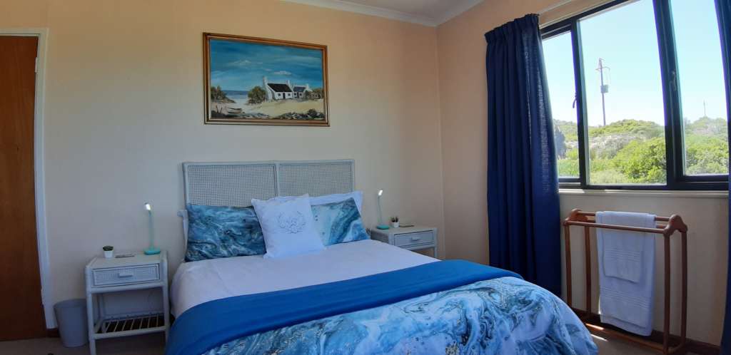 Bedroom 1 at Fonteintjies. There is a double bed with a white headboard. Above the bed is an oil painting of a seaside cottage. There are bedside tables with lamps at both the left and right of the bed. There is a window in the wall to the right, and a door in the far left corner.