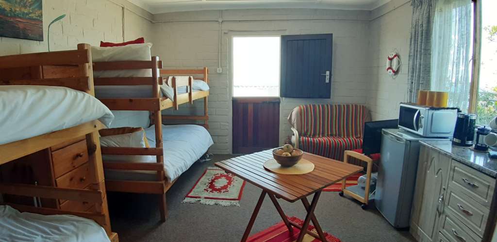 The annex room at Fonteintjies. In the centre foreground is a square table with a bowl of fruit. On the right are a kitchen cupboard with a kettle and mugs on top, and a small fridge with a microwave oven on top. In the corner to the far right is a couch draped with a striped blanket, with a TV facing the couch. In the centre rear is a door open to the outside. Along the wall to the left are four bunk beds.