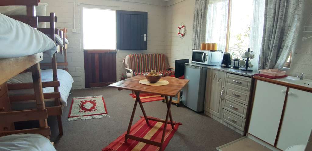 The annex room at Fonteintjies. In the centre foreground is a square table with a bowl of fruit. On the right near the foreground are a kitchen sink, kitchen cupboard with a kettle and mugs on top, and a small fridge with a microwave oven on top. In the corner to the far right is a couch draped with a striped blanket, with a TV facing the couch. In the centre rear is a door open to the outside. Along the wall to the left are four bunk beds.