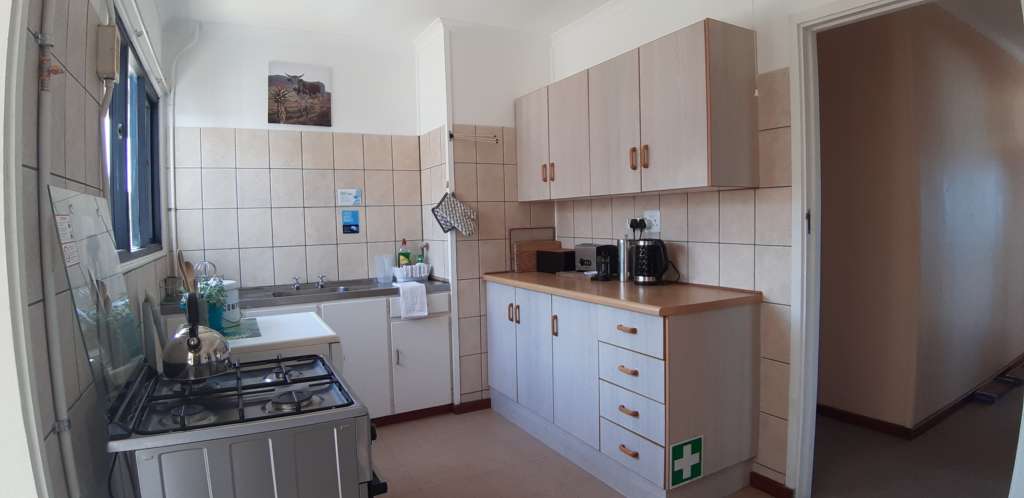The kitchen at Fonteintjies. There is a gas stove with a kettle on the hob, a dishwasher just visible behind the stove, a sink, and a cupboard unit. On the counter are a bread bin, toaster, kettle, and coffee plunger. A first aid sign appears near the bottom drawer.