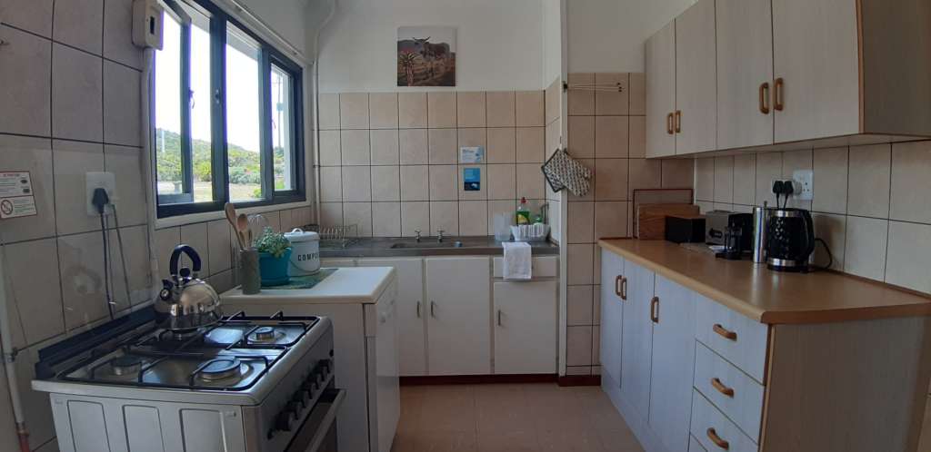 A wide view of the kitchen at Fonteintjies. On the left are a gas stove with a kettle on the hob, and a dishwasher with a cooking utensils, a pot plant, and a compost bucket on top. In the centre is a sink. On the right is a cupboard and counter. On the counter are cutting boards, a bread bin, a toaster, a kettle, and a coffee plunger.
