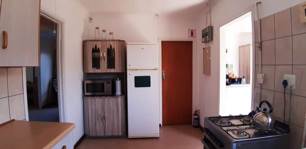 The kitchen at Fonteintjies. At the left is the end of a counter, and a doorway to the dining room. In the centre rear is a cupboard unit with a microwave oven on the counter. To its right is a white two-door fridge/freezer. Next to a closed door is a fire extinguisher. On the right is a silver gas stove with a kettle on the hob. Behind the stove is a doorway to the entrance portal.
