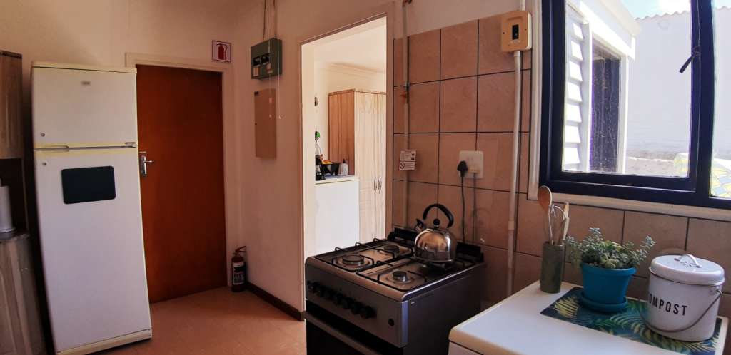 The kitchen at Fonteintjies. There is a white two-door fridge/freezer, a fire extinguisher, a silver gas stove with a kettle on the hob, and the white top of a dishwasher. On top of the dishwasher are a container of cooking utensils, a pot plant, and a bucket labeled "compost". A doorway leads to the entrance portal where a cupboard and a small fridge are just visible.