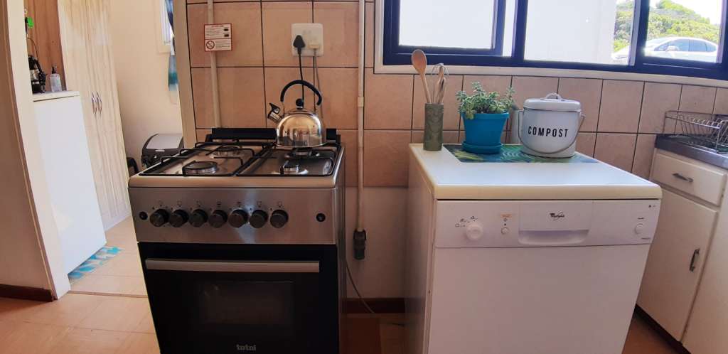 The kitchen at Fonteintjies. In the centre left is a silver gas stove with a kettle on the hob. In the centre right is a dishwasher, with cooking utensils, a pot plant, and a bucket for collecting compostable waste.