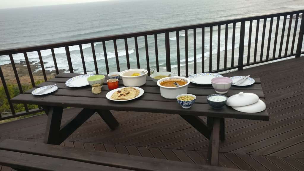 On the deck at Fonteintjies is a wooden picnic bench, laid out with food for a sunset dinner overlooking  the ocean. Just behind the table is the deck railing, and beyond that is a view of the ocean.