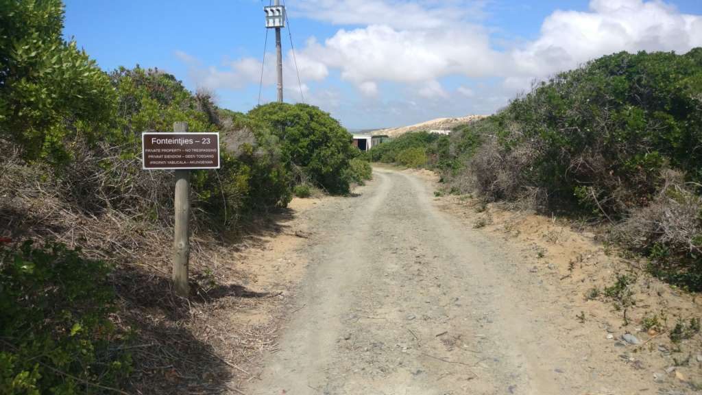 The entrance road at Fonteintjies. It's a gravel road with green vegetation to the left and right. In the left foreground is a sign with the name "Fonteintjies", the erf number, and "Private Property - No Trespassing" in English, Afrikaans, and Xhosa. In the far distance you can see parts of some outbuildings. In the middle distance to the left is a pole with an electrical transformer.
