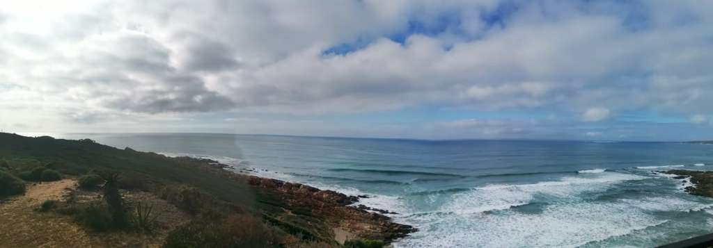A panoramic view of the ocean and a rocky shore, seen from the deck at Fonteintjies. an aloe is in the foreground to the left. Two neighbouring houses are in the distance to the left.