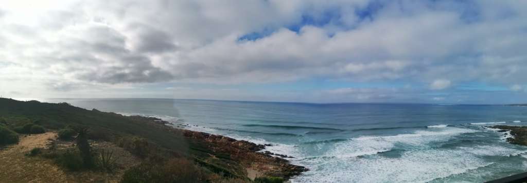 Ein Panoramablick auf das Meer und ein felsiges Ufer, vom Deck von Fonteintjies aus gesehen. eine Aloe ist im Vordergrund links. Zwei benachbarte Häuser sind in der Ferne auf der linken Seite.